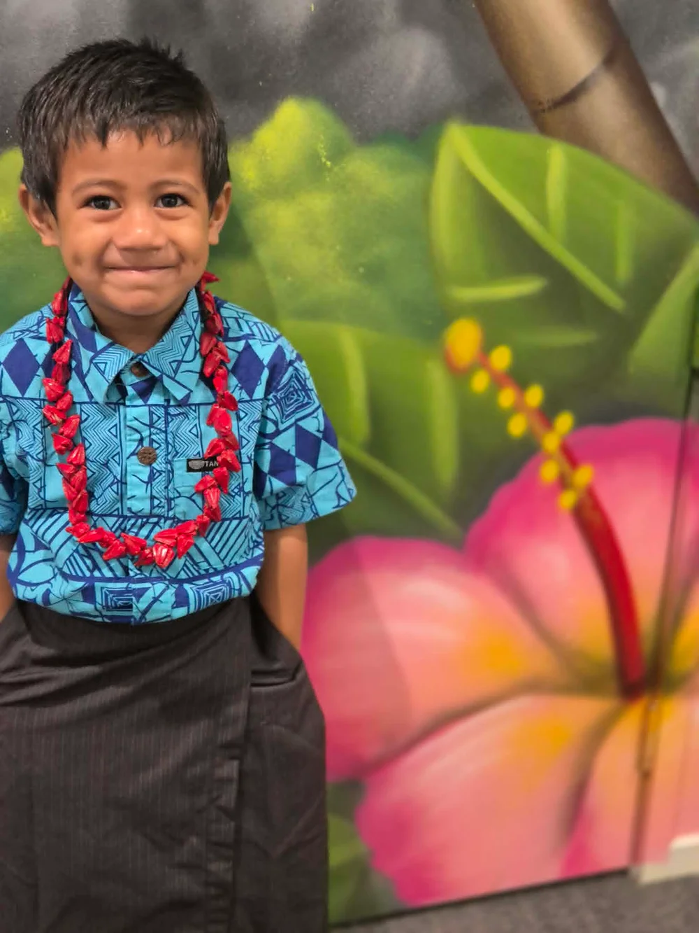 A Tiny Stars tamaiti wearing a flower lei and blue patterned shirt